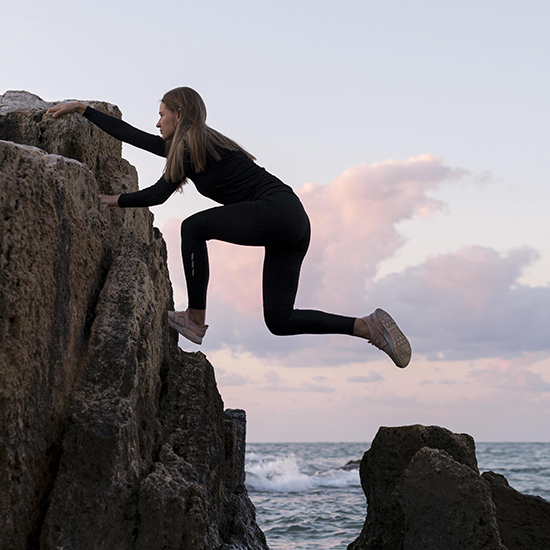 A woman dressed in black athletic clothing is climbing a large rock formation by the ocean. Her right hand and foot are gripping the rock as she reaches upward with her left hand. The sky is partly cloudy with the sea visible in the background.