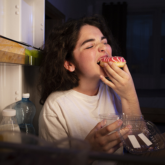 A person with dark, curly hair is seen enjoying a glazed doughnut with pink icing. They are standing in front of an open refrigerator, holding a transparent container. The scene appears to be at night, lit by the refrigerator's light.