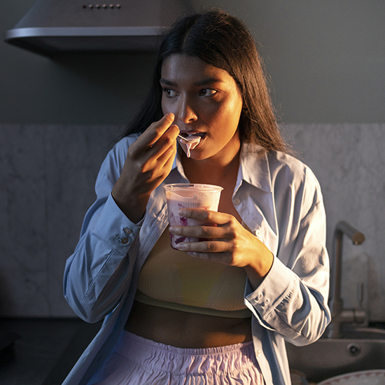 A woman with long dark hair is standing in a kitchen, eating from a yogurt cup. She is wearing a light blue shirt unbuttoned over a yellow top and pink bottoms. The lighting suggests it's either evening or early morning. She appears thoughtful while eating.