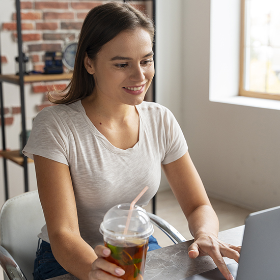 A woman in a white t-shirt is sitting at a table in a room with a brick wall, holding a plastic cup with a straw and looking at her laptop screen. She appears to be smiling and engaged with what is on the screen.