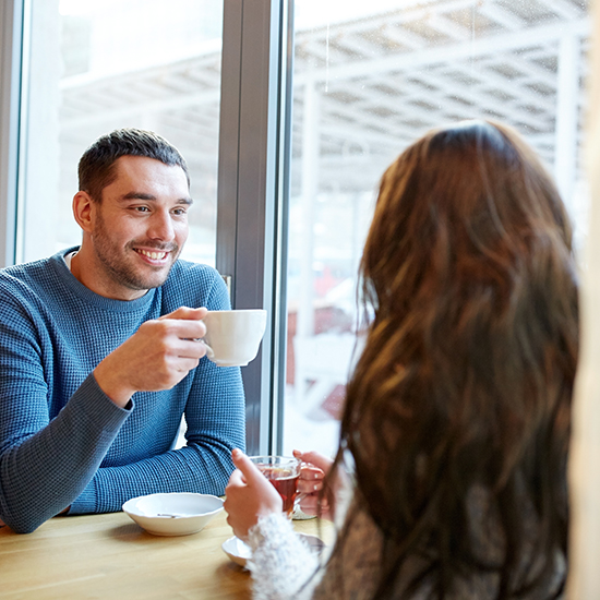 A man and woman sit at a table by a window, engaged in conversation. The man, wearing a blue sweater, holds a coffee cup and smiles. The woman, seen from behind, has long brunette hair and wears a sweater, also holding a cup. The setting is a bright cafe.