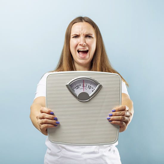 A woman with long brown hair and a white shirt holds a bathroom scale towards the camera. She has an open-mouthed, frustrated expression. The background is light blue.