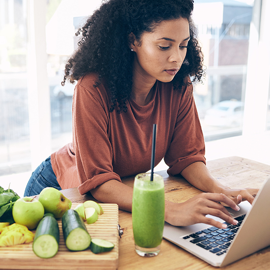 A woman with curly hair, focused and typing on a laptop at a wooden table. On the table is a cutting board with cut cucumbers, apples, and other fruits and vegetables, and a glass of green smoothie with a straw. Light streams in from nearby windows.