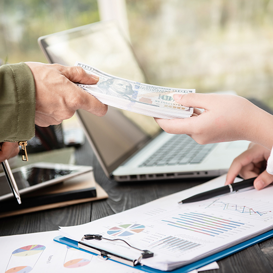 A person hands a stack of dollar bills to another person over a table with financial charts and graphs, calculator, tablet, and laptop.