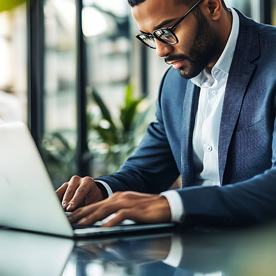 A man wearing black-framed glasses and a blue blazer is working on a laptop at a desk. The background features large windows and indoor plants, giving the space a modern and bright atmosphere.