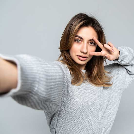 A woman with shoulder-length light brown hair is taking a selfie. She is wearing a light gray sweater and making a peace sign with her right hand. The background is plain gray.