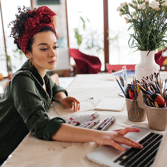 A woman with a red headscarf and curly hair works on a laptop at a table filled with art supplies, including markers, paintbrushes, and a vase of flowers. She appears focused, with a notebook also visible on the table.