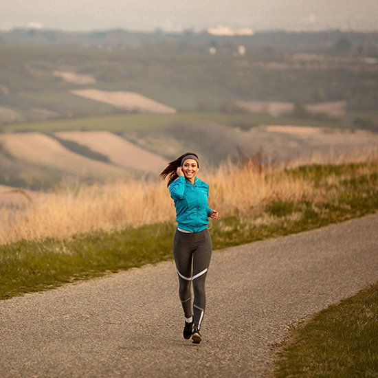 A woman is jogging on a paved path through a hilly, grassy landscape. She is wearing a turquoise jacket, black leggings, and black running shoes, and she has a headband on. She appears to be smiling and holding a smartphone to her ear. The sky is overcast.