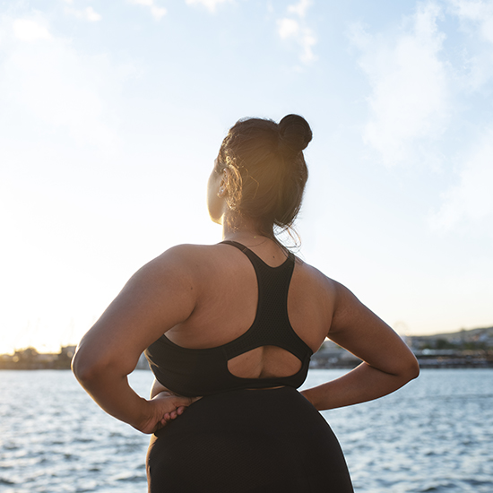 A person stands with their back to the camera, hands on hips, looking out over a body of water. They are dressed in a black sports top and have their hair tied up in a bun. The sun is setting, casting a warm glow over the scene.