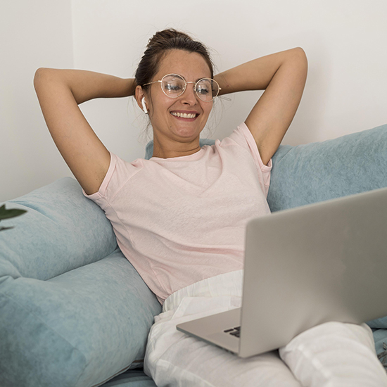 A woman with glasses and earbuds relaxes on a blue couch with her hands behind her head, smiling while looking at her laptop. She wears a light pink t-shirt and white pants.