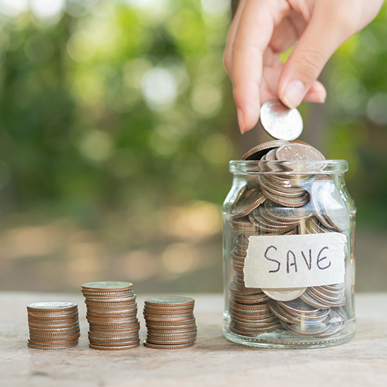 A hand places a coin into a glass jar labeled "SAVE," which is filled with coins. Three neatly stacked piles of coins are arranged next to the jar on a wooden surface. The background is blurred greenery.