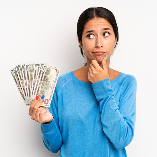 A person with a thoughtful expression holds several US dollar bills fanned out in one hand. They are wearing a blue sweater and have their other hand touching their chin. The background is plain white.