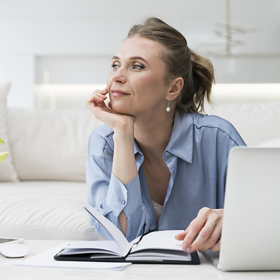 A woman with a thoughtful expression sits at a table, her elbow resting on the table and her hand on her face. She wears a light blue shirt and has a notebook open in front of her, with a laptop to her side. The background shows a well-lit, modern interior.