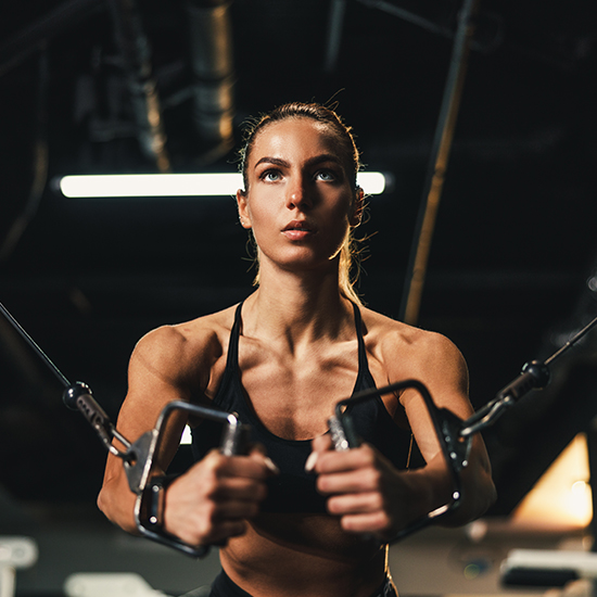 A woman in a gym performs a chest workout using cable machines. She is focused, wearing a black sports bra, with her hair pulled back. The background is dimly lit, emphasizing the physical effort and concentration.