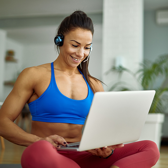 A woman wearing a blue sports bra and headphones smiles while looking at a laptop. She is sitting cross-legged in a bright room with plants and home furnishings in the background.