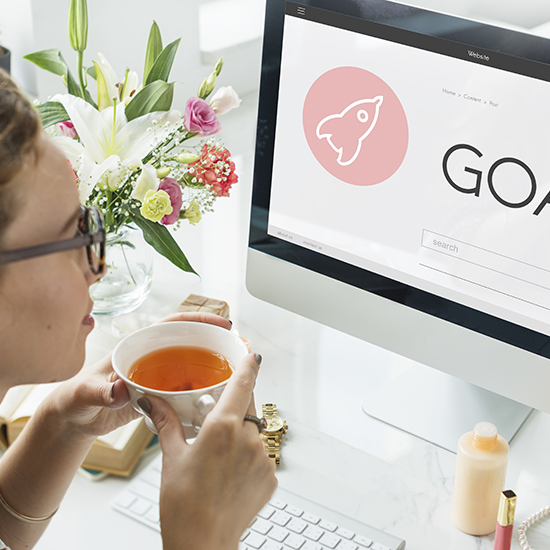 A person in glasses drinks tea while looking at a computer screen displaying a webpage with a rocket icon and the word "GOALS." The desk has a bouquet of flowers, an open book, a smartphone, and some cosmetics.