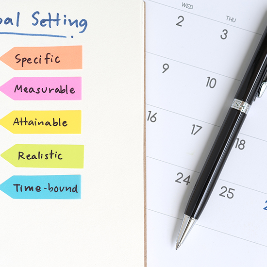 A notebook page titled "Goal Setting" lists five colorful sticky notes with the words: Specific, Measurable, Attainable, Realistic, and Time-bound. Beside the notebook, there is a calendar with a black pen placed on it, showing mid-month dates.