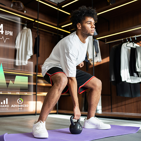 A man in a white shirt and black shorts performs a kettlebell exercise on a purple mat in a modern home gym. He is holding a black kettlebell in a squat position. Digital overlays show workout intensity, duration, and heart rate. Clothing is displayed in the background.