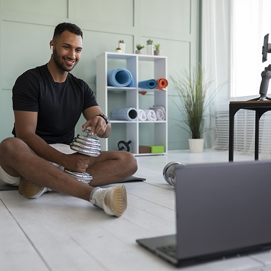 A man sits cross-legged on the floor in front of a laptop, holding a stack of silver dumbbells. He is in a bright, modern room with neatly arranged exercise equipment on shelves behind him. He is wearing a black T-shirt and white shorts and smiling at the laptop screen.