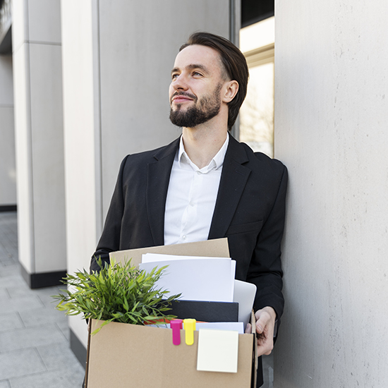 A man with a beard and wearing a black blazer and white shirt leans against a wall outdoors. He is holding a cardboard box filled with office supplies, a small plant, binders, and a sticky note. He is looking upward with a slight smile.