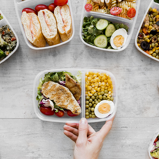 A variety of meal prep containers are displayed on a light wooden surface. These include a sandwich with tomatoes, a salad with cucumbers and eggs, and a container with grilled chicken, peas, corn, and a boiled egg. A hand is holding one of the containers.