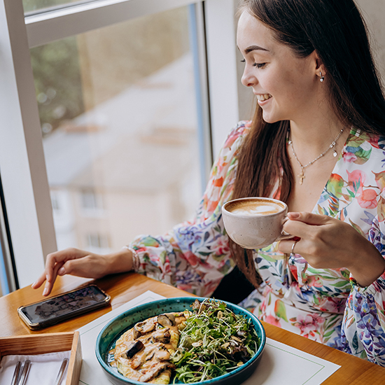 A woman with long brown hair sits by the window at a restaurant. She is holding a cup of coffee and smiling while looking at her smartphone on the table. In front of her is a blue bowl filled with various greens and vegetables. She wears a colorful floral top.