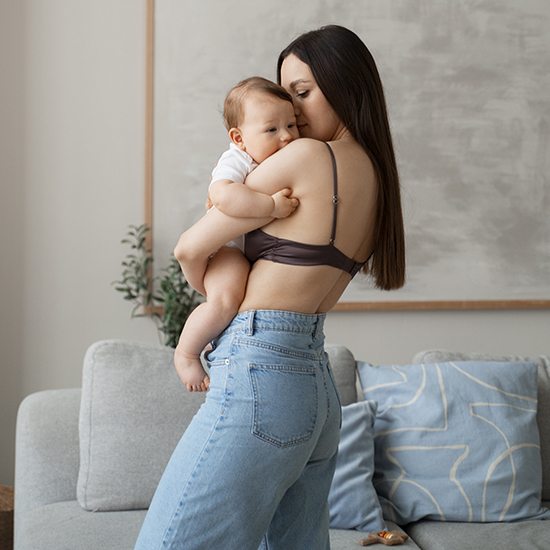 A woman with long, dark hair is holding a baby in her arms. She is wearing a dark crop top and light blue jeans. The baby is dressed in a white onesie and has light brown hair. They are standing in a room with a gray couch and a plant in the background.