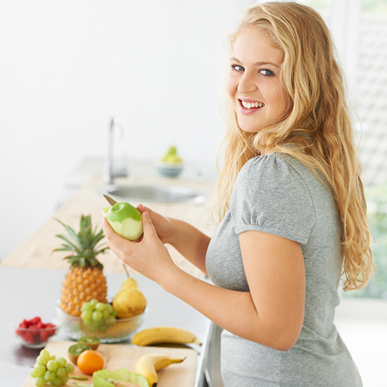 A smiling woman with long blonde hair stands in a kitchen, holding a green apple. On the counter in front of her, there is an assortment of fresh fruits, including pineapples, bananas, grapes, and pears. She is wearing a gray shirt.