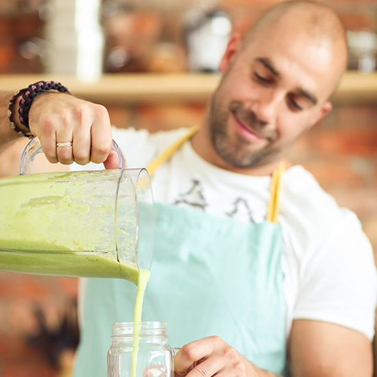 A bald man with a beard wearing a turquoise apron and a white t-shirt is smiling while pouring a green smoothie from a blender into a mason jar. The background is slightly blurred with warm tones, suggesting a cozy kitchen setting.