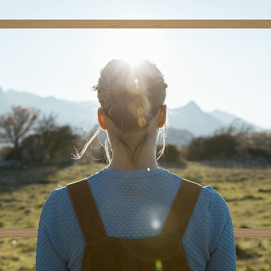 A person with light hair in a bun, wearing a blue sweater and brown overalls, faces a mountainous landscape. The sun shines directly above their head, partially covered by their hair. The foreground is a grassy field with scattered trees.