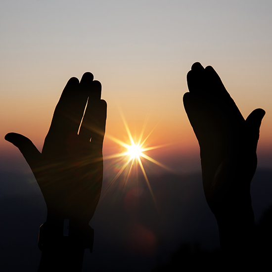 Silhouetted hands are raised in the air against a backdrop of a stunning sunset, with the sun positioned between the hands, casting a radiant starburst effect. The sky has a gradient of warm colors from orange to purple.