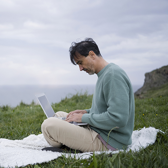 A man with dark hair, wearing a green sweater and beige pants, sits cross-legged on a white blanket on a grassy hill. He is working on a laptop with a cloudy sky and rocky landscape in the background.