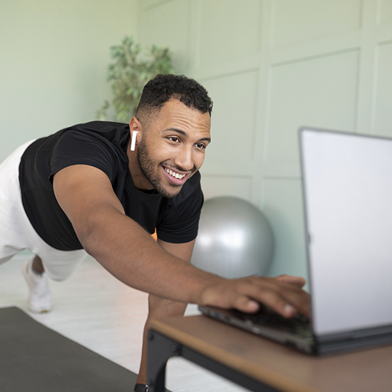 A man in workout attire smiles as he follows an online fitness class on his laptop. He is doing a plank exercise on a yoga mat, with one hand tapping the laptop. He is wearing wireless earbuds, and a large exercise ball is in the background.