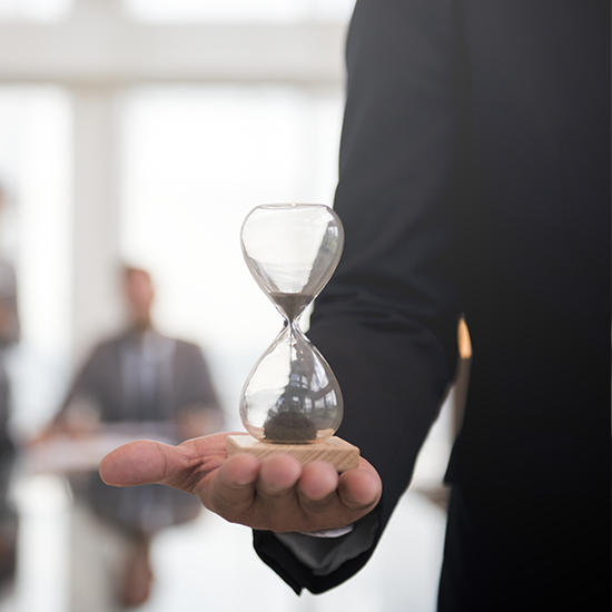 A person in a suit holds a wooden hourglass with sand flowing through it, indicating the passage of time. Blurred background features two other people in a modern office setting. The image conveys a sense of urgency or time management.