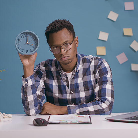 Man with glasses and a checkered shirt holding up a blue clock, looking serious. He is seated at a desk with paper, a pen, and a laptop. Blue background with sticky notes.