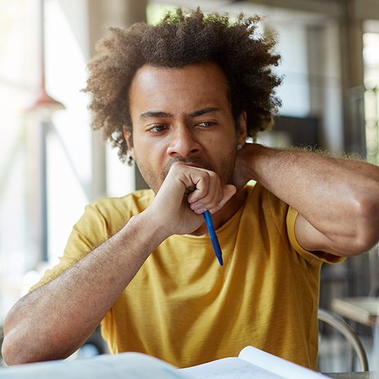 A person sits at a table with an open book in front of them, holding a pen in one hand and resting their head on the other hand. They appear deep in thought, with a slightly furrowed brow. The background is blurred, suggesting an indoor setting.