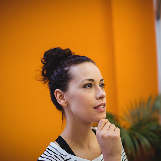 A person with dark hair tied in a bun is gazing thoughtfully to the side against a vibrant orange background. They are wearing a white and black striped top and resting their chin on their hand. There is a green plant visible in the background.