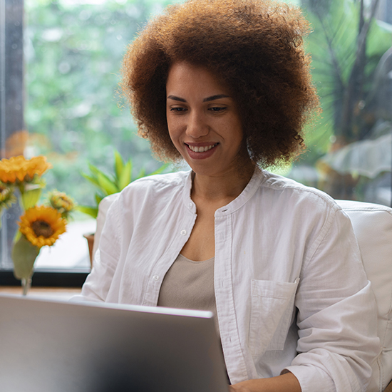 A person with curly hair smiles while looking at a laptop screen. They are wearing a white shirt and sitting in a bright room with green plants and yellow sunflowers in the background. Natural light filters through a large window.