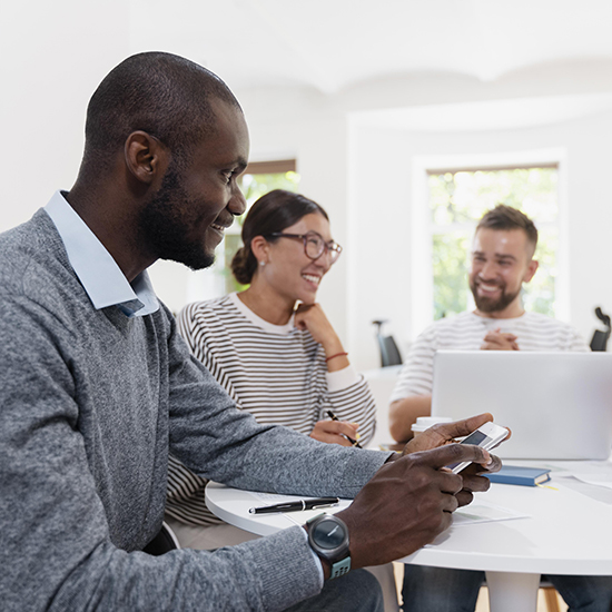 A group of three people sit around a table, having a discussion. The person in the foreground is holding a smartphone and smiling. The other two, one wearing glasses and the other with a beard, are also smiling, with a laptop in front of them.