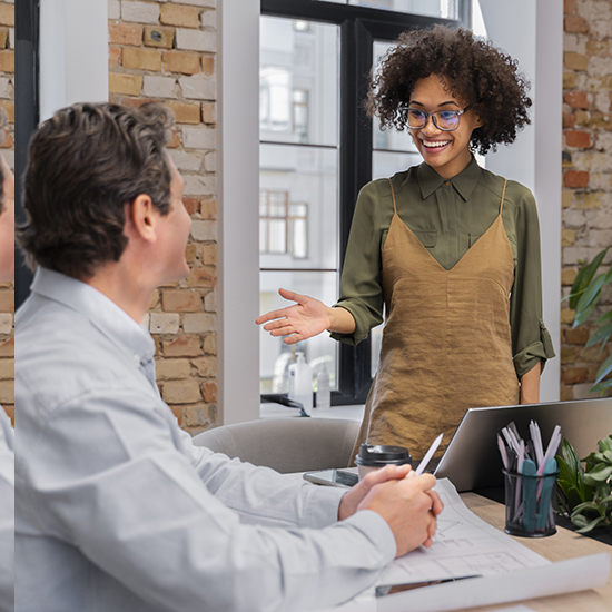 A woman with curly hair and glasses, wearing a green shirt and beige dress, stands in an office smiling and gesturing while speaking to a seated man. They are in a modern office with exposed brick walls, large windows, and laptops on the table.