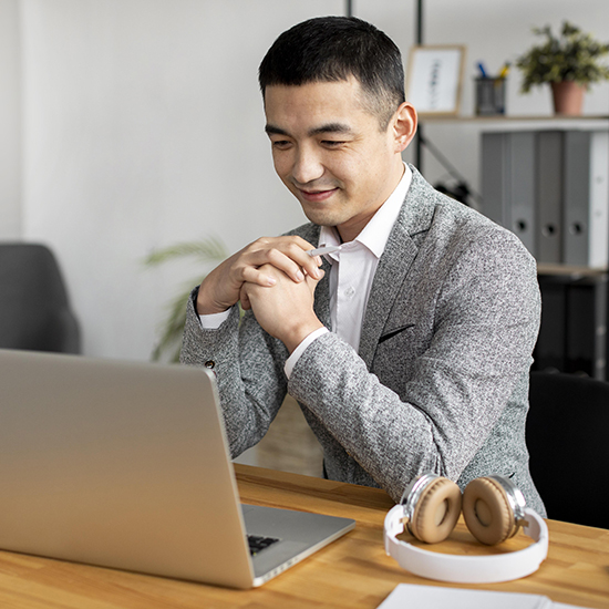 A man in a grey blazer and white shirt is smiling and looking at a laptop on a wooden desk. He has his hands folded in front of him. A pair of headphones is sitting on the desk beside the laptop. Shelves and plants are in the background.