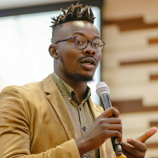 A man with short, twisted hair and glasses is holding a microphone and speaking. He is wearing a brown blazer over a patterned shirt and appears to be presenting or addressing an audience in an indoor setting. The background shows a blurred modern interior.