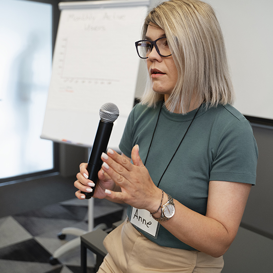 A person with glasses and shoulder-length blonde hair holds a microphone while speaking. They are wearing a green shirt, beige pants, and have a name tag reading "Anne." A flip chart with a graph labeled "Monthly Active Users" is visible in the background.