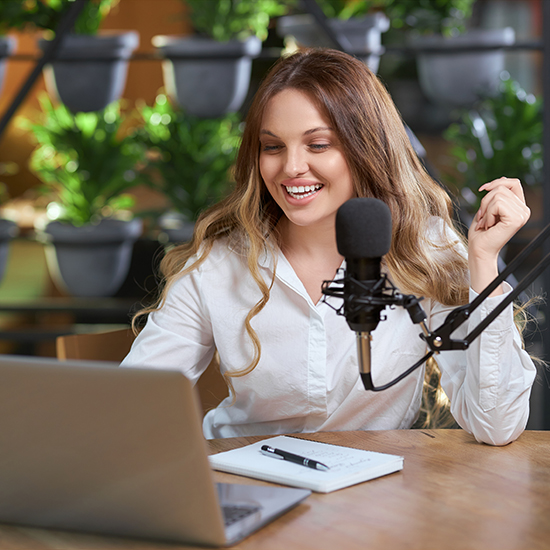 A smiling woman with long hair sits at a wooden desk with a laptop open in front of her. She wears a white shirt and speaks into a microphone on a stand. A notebook and pen are on the table. There are potted plants on shelves in the background.