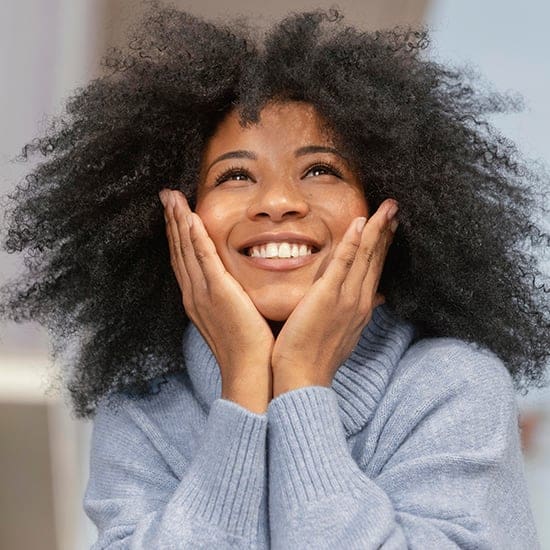 A woman with dark curly hair and a bright smile rests her chin on her hands, looking upward. She is wearing a light blue turtleneck sweater, and the background appears to be softly lit. The image conveys a cheerful and hopeful expression.