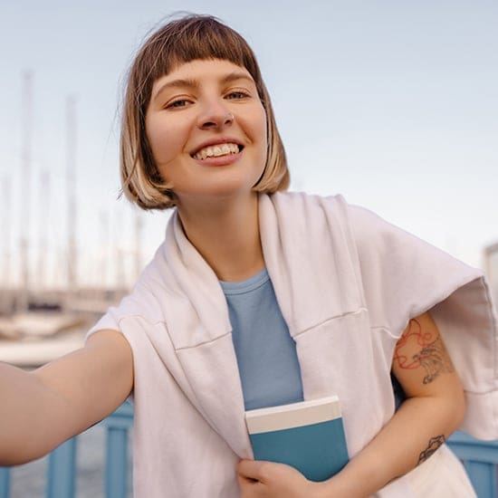 A young woman with a short bob haircut smiles while taking a selfie outdoors. She has a white sweater draped over her shoulders and holds a blue book. In the background, there are boats docked at a marina.