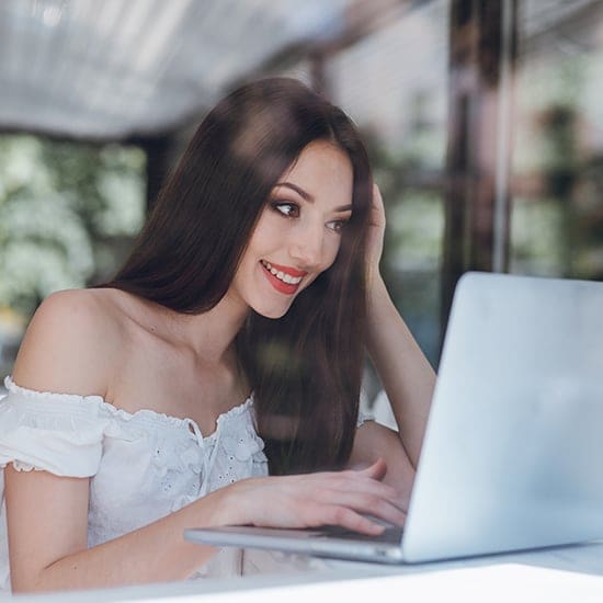 A woman with long brown hair and a white off-shoulder top is smiling and looking at a laptop screen. She is sitting in a bright, airy room with large windows and greenery visible outside.