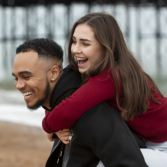 A smiling young woman in a red sweater rides piggyback on a laughing young man with a short beard. They are outdoors near a beach, with an out-of-focus pier in the background. Both appear to be enjoying each other's company.