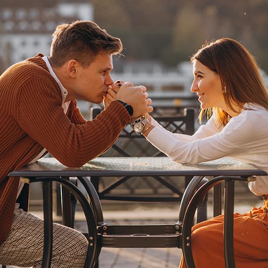 A couple sits at an outdoor café table, holding hands and looking intently at each other. The man, wearing a brown sweater, kisses the woman's hand, while she, dressed in a white top, gazes at him with a smile.