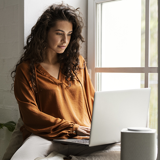 A woman with long curly hair, wearing an orange blouse, is sitting on a bed by a window using a laptop. She appears focused, with natural light illuminating the room. An out-of-focus object is in the foreground.